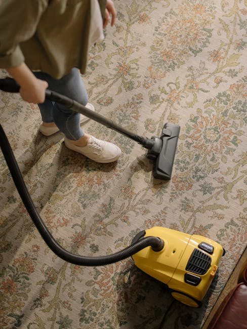 A person using a vacuum cleaner with a yellow body and black hose attachment to clean a patterned, beige-colored carpet in a residential living room. The individual is wearing beige shoes, blue jeans,