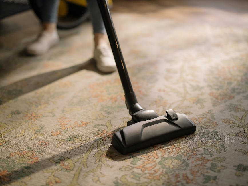 Close-up of a black vacuum cleaner head actively cleaning a patterned, beige and green carpet in a residential setting. The vacuum cleaner's hose and part of the handle are visible, with the cleaning 