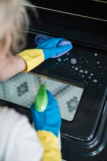 A professional cleaner wearing yellow and blue rubber gloves is cleaning the glass surface of a black oven door using a cloth and a spray bottle. The oven's interior is visible through the glass, show
