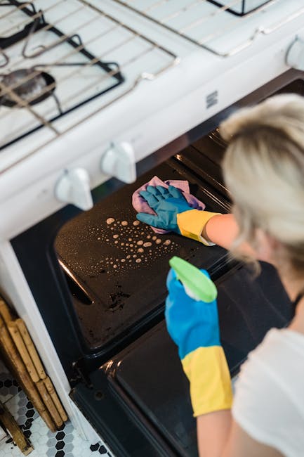 A professional cleaner wearing yellow and blue rubber gloves is cleaning the glass surface of a black oven door using a cloth and a spray bottle. The oven's interior is visible through the glass, show