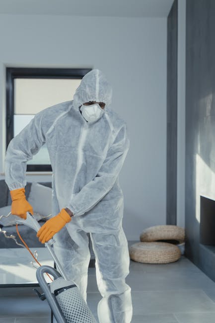 A professional cleaner dressed in white protective coveralls, a face mask, and orange gloves is performing surface cleaning with a handheld device in a modern residential room. The room features a smooth, light-colored tiled floor, a large window allowing natural light, and minimal decor including a pair of round, beige cushions placed on the floor. The cleaner appears to be thoroughly sanitising the tiled surface, ensuring hygiene and cleanliness. Visible sunlight creates bright, shadowed areas on the surfaces, reflecting the high standard of deep cleaning services provided by Cleaners Holland Park, especially suitable for preparing homes after events or for regular maintenance. The scene emphasizes meticulous high-touch surface sanitation in a contemporary living space, demonstrating their expertise in domestic cleaning.